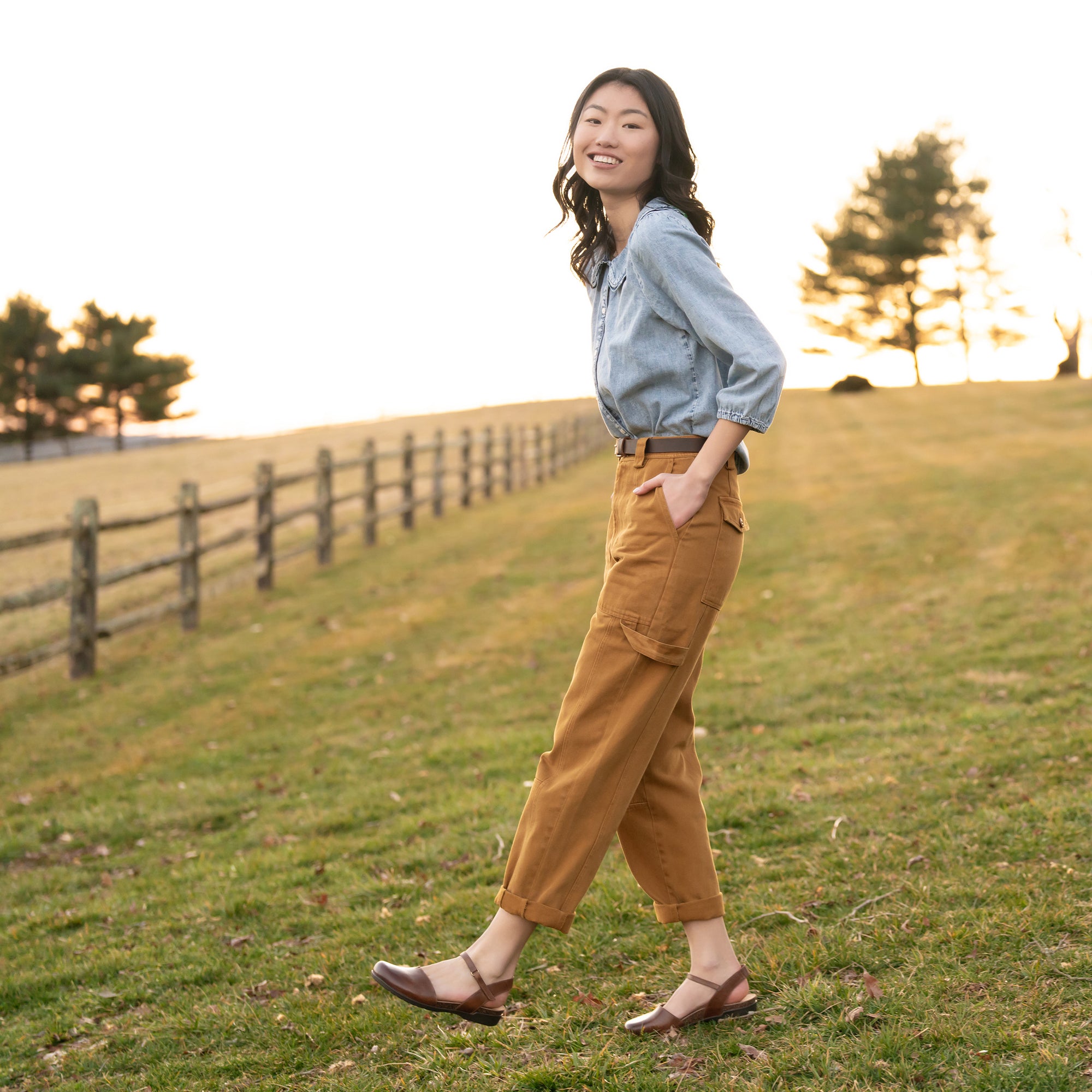 A woman standing in a field wearing brown waxy leather closed-toe sandals.