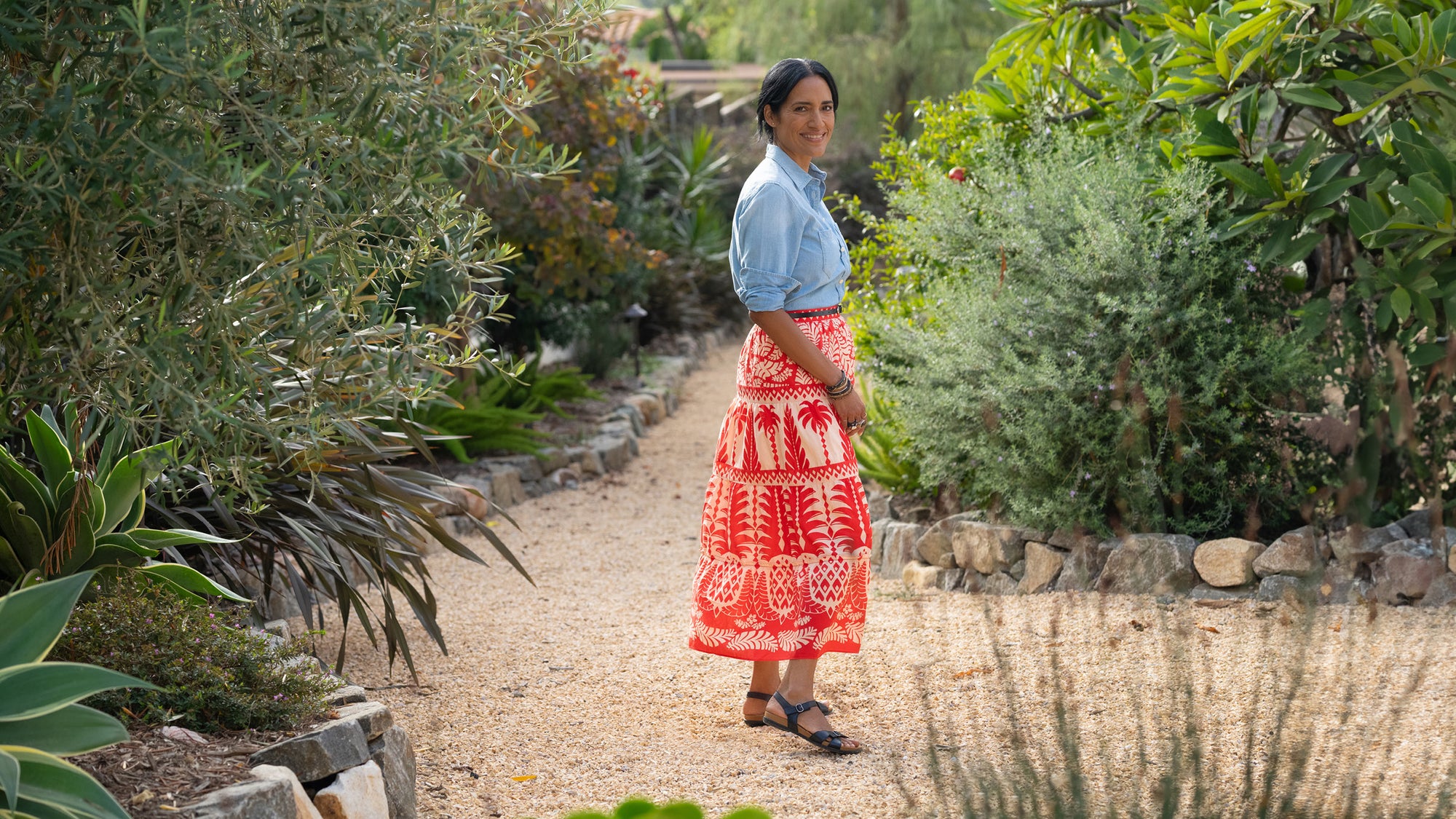 A woman standing in a garden in a bright skirt and black flat sandals.