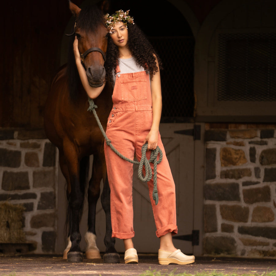 Woman with horse in stylish overalls wearing light tan clogs with supportive outsoles.
