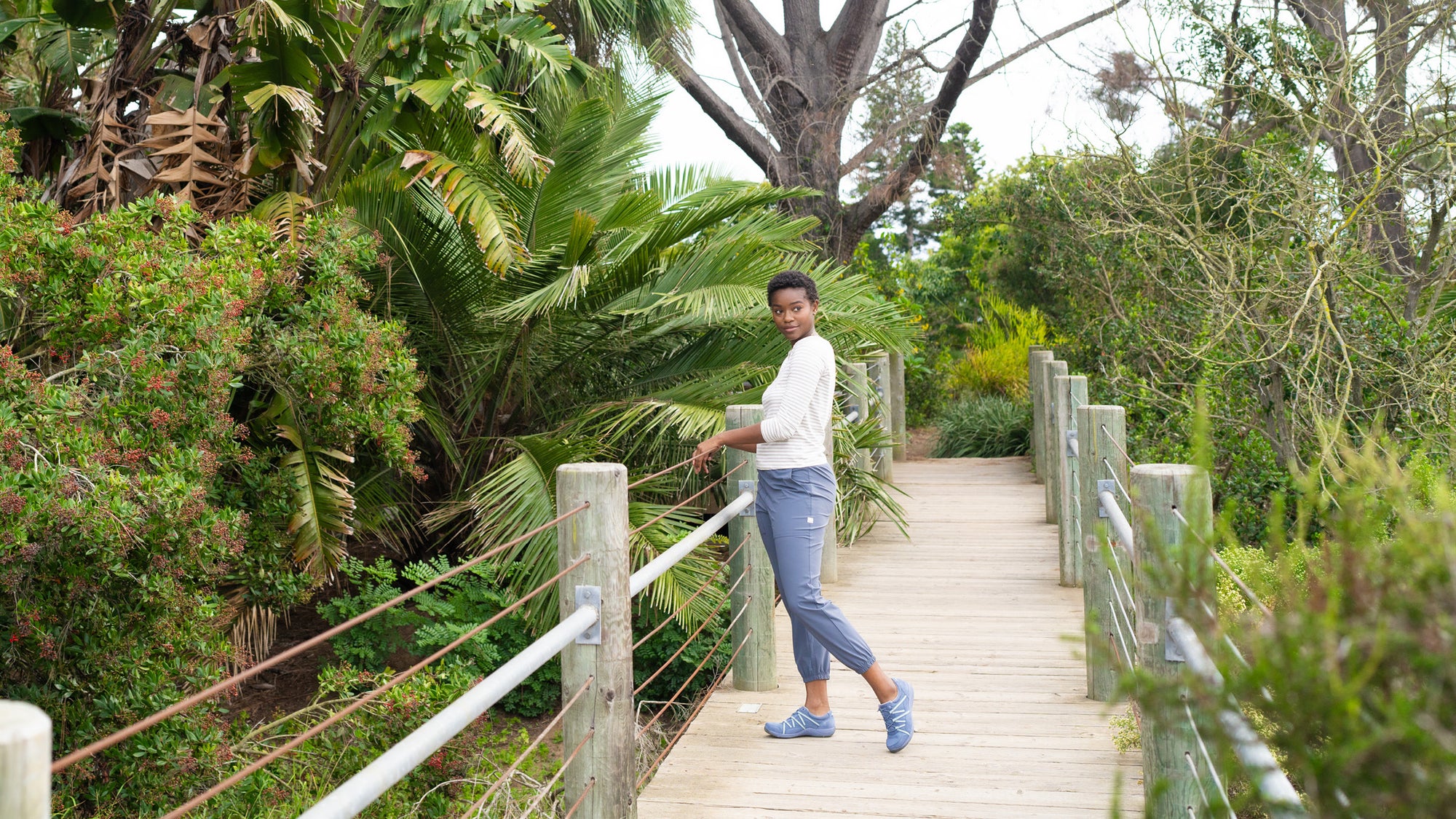 A woman standing on a bridge in nature wearing comfortable attire and lightweight blue sneakers.