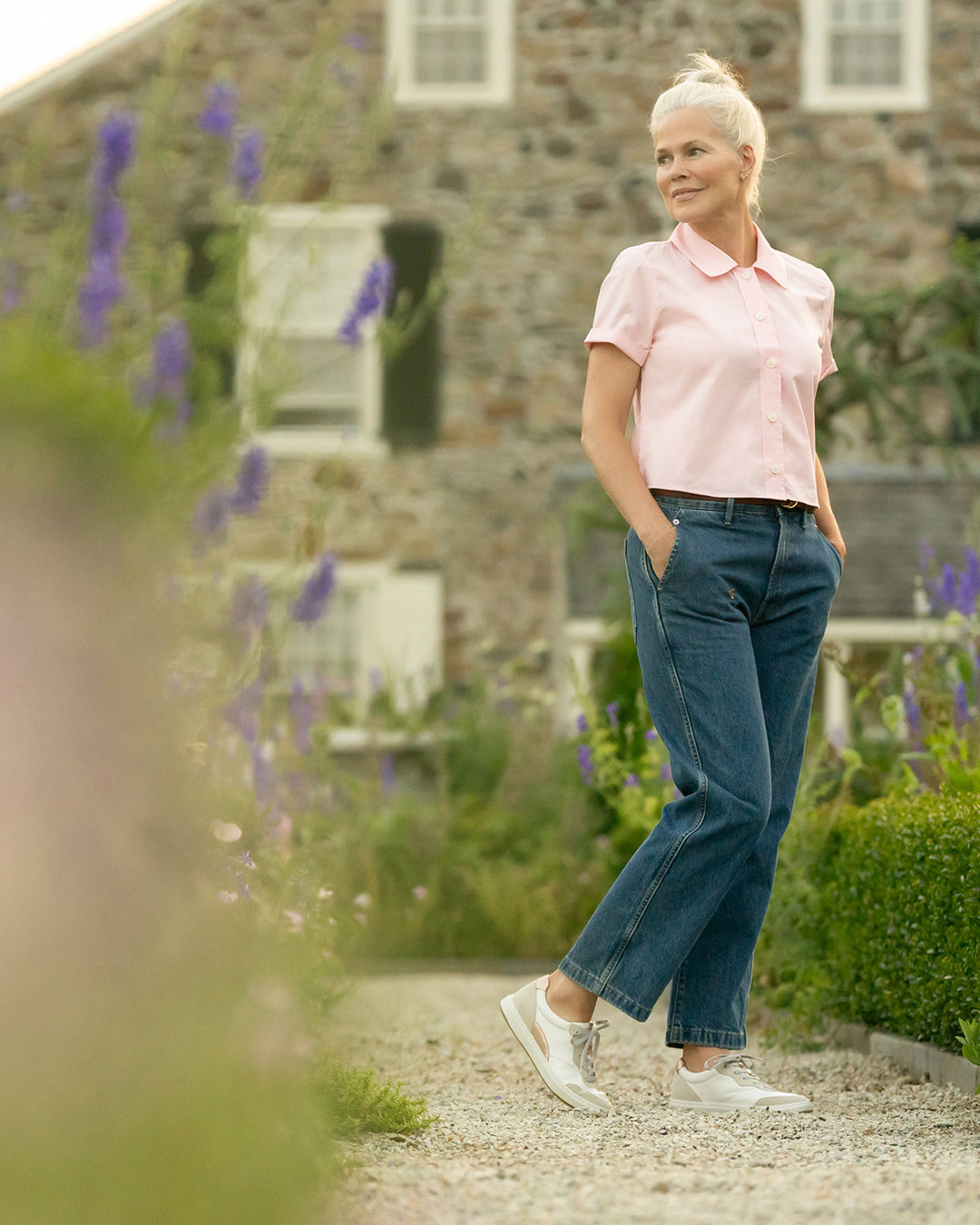 Woman in pink blouse, blue jeans and white and rose gold sneakers in outdoor garden.