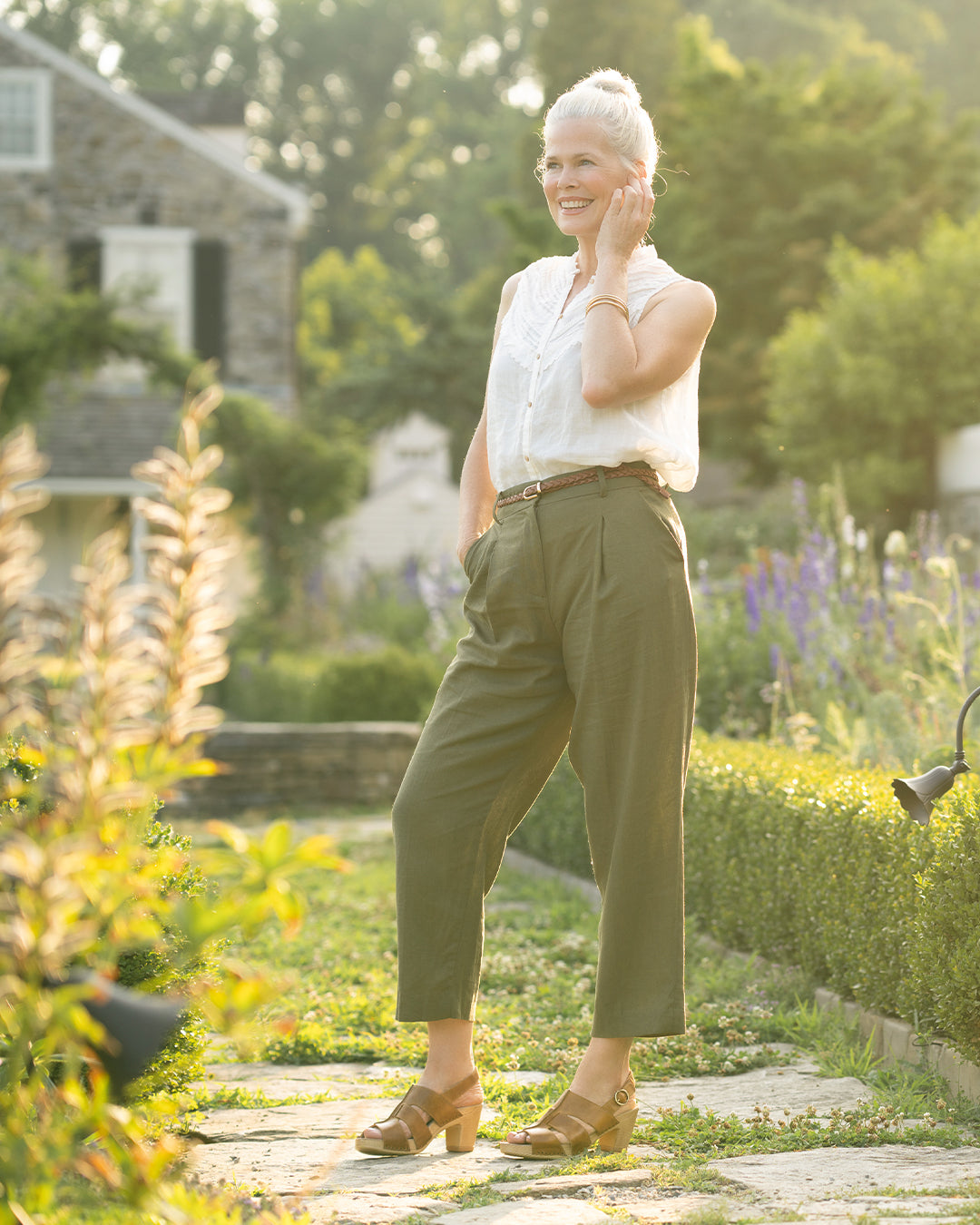 Woman in outdoor garden in white blouse and green pants with tan heeled sandals.