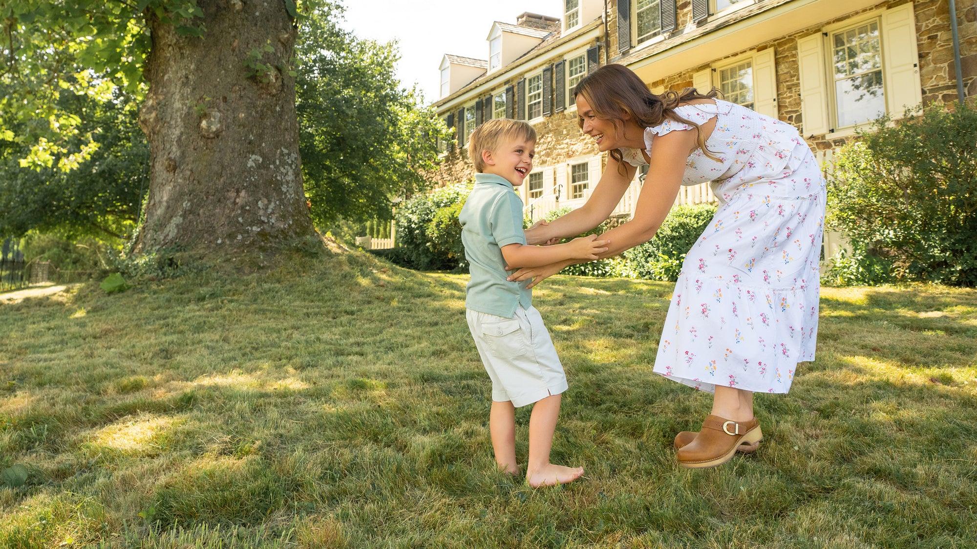 Woman standing with her child in front of stone house. Woman is wearing floral dress and tan Dansko mules. Child is wearing a polo and khaki shorts.