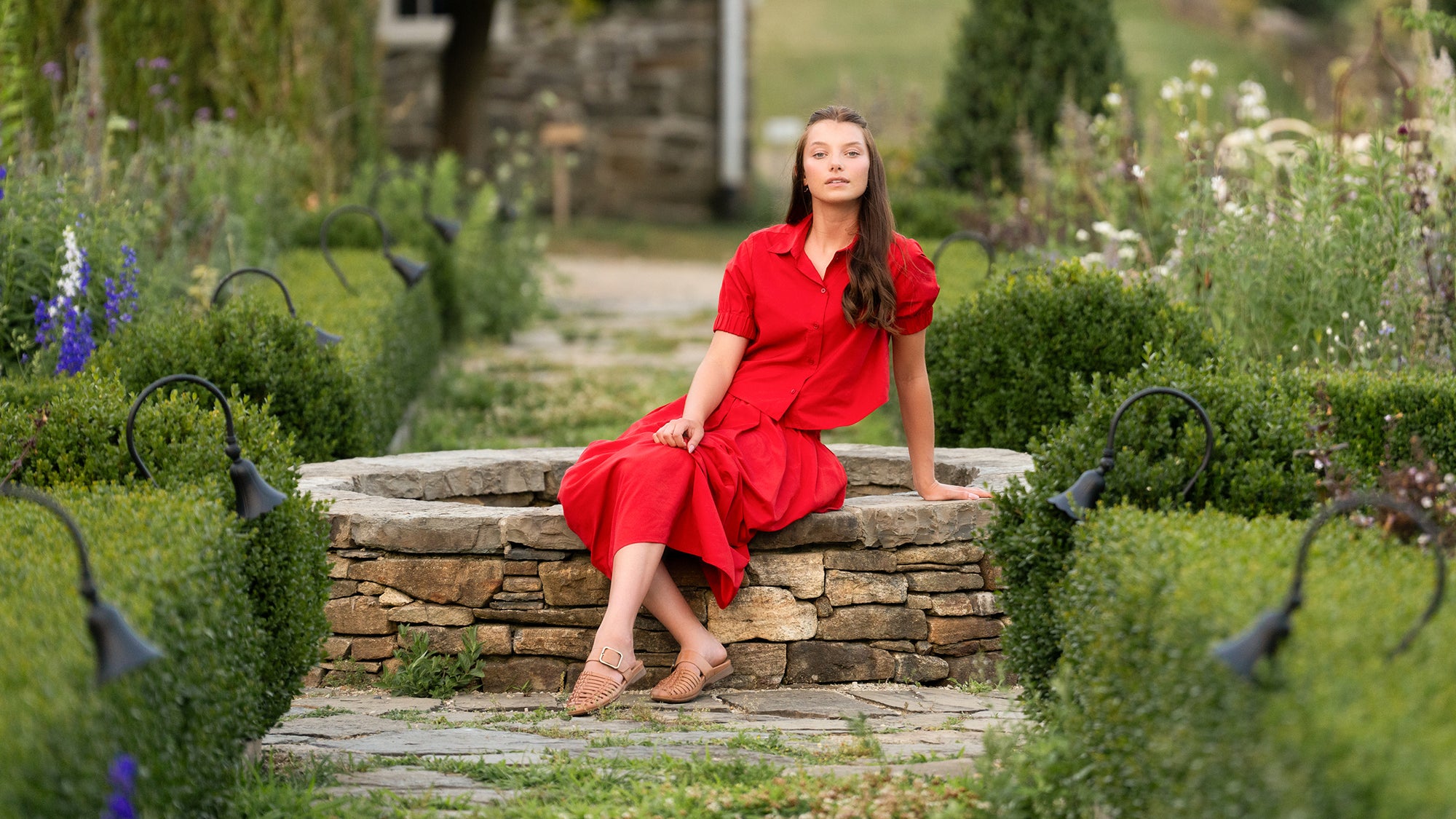 girl in red shirt and skirt sitting on stone fountain in tan sandals.