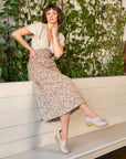 Woman in a floral skirt and light shoes leaning against a white wall with greenery.