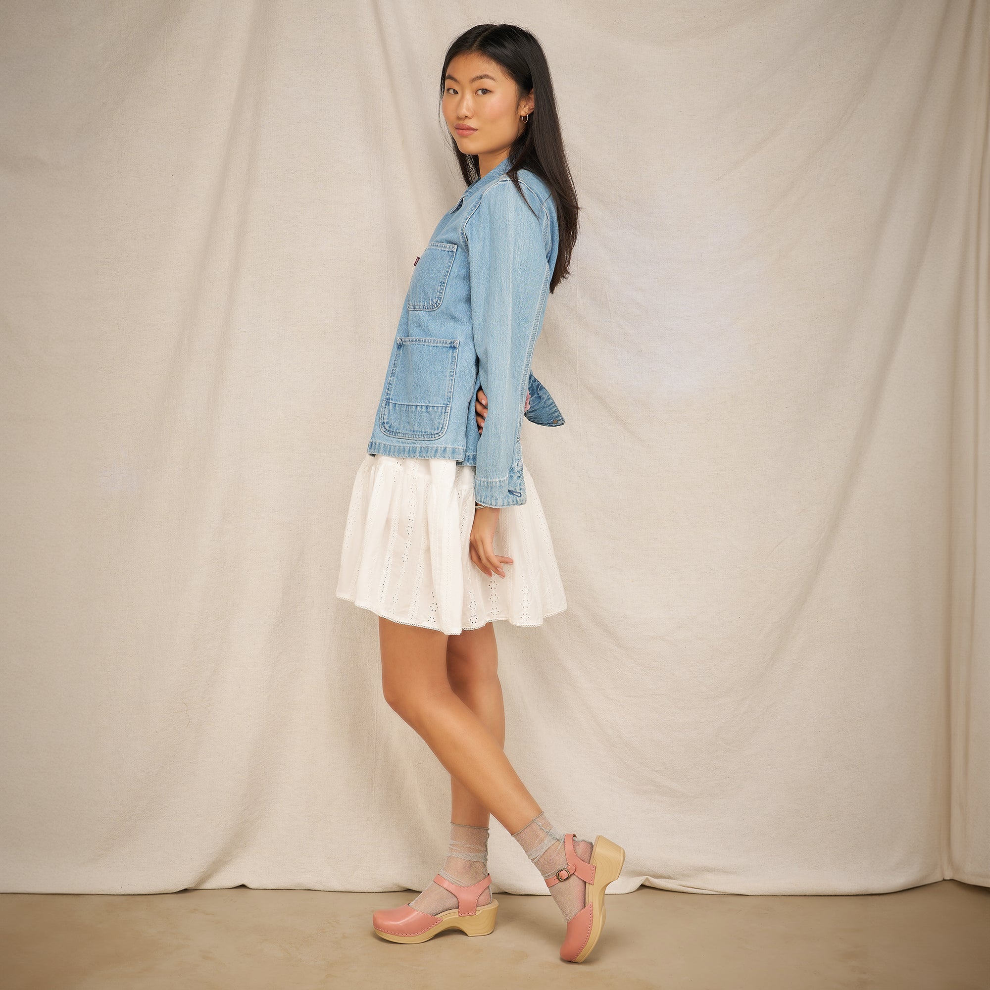 A woman standing against a plain backdrop while wearing pale pink clog sandals.