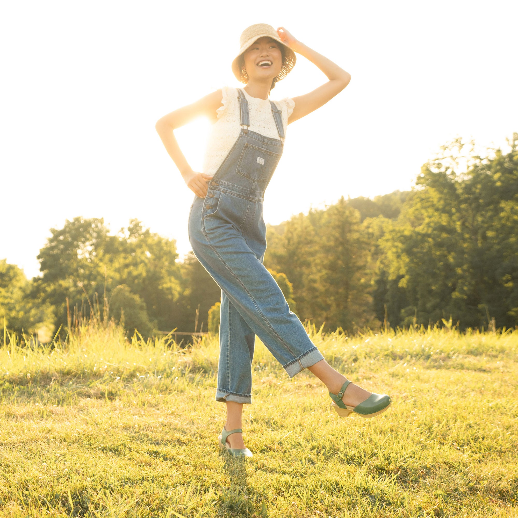 A woman in a field wearing overalls and pale green flatform sandals.