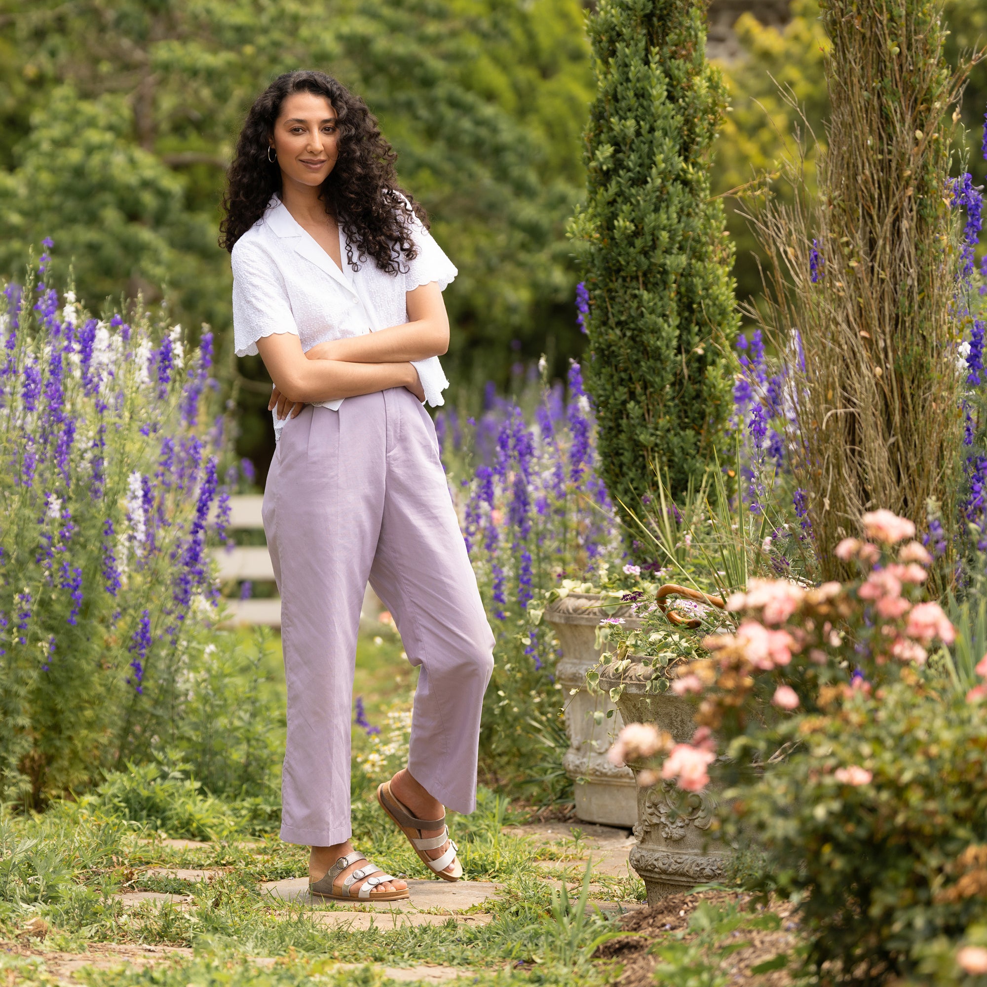 Woman standing in garden wearing white blouse, lavender trousers, and multi-colored metallic sandals.