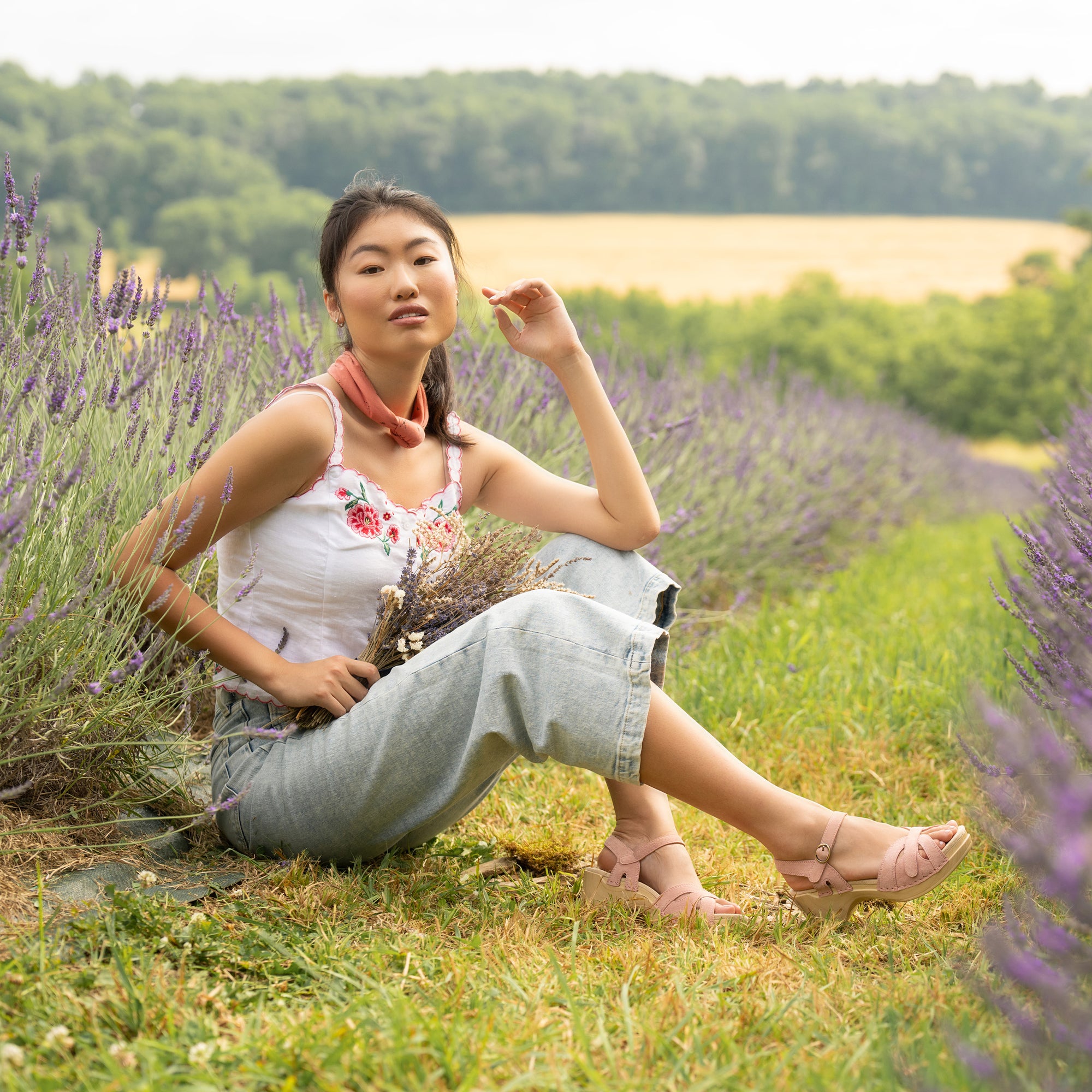 Woman sitting in lavender field wearing a white tank top with floral detail, cropped jeans and pink Dansko sandals.