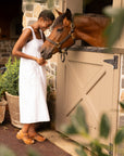 A woman in a white dress standing with a horse while wearing tan flatform sandals.