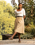 A woman standing in a garden in a white top and tan skirt while wearing black two-strap sandals.