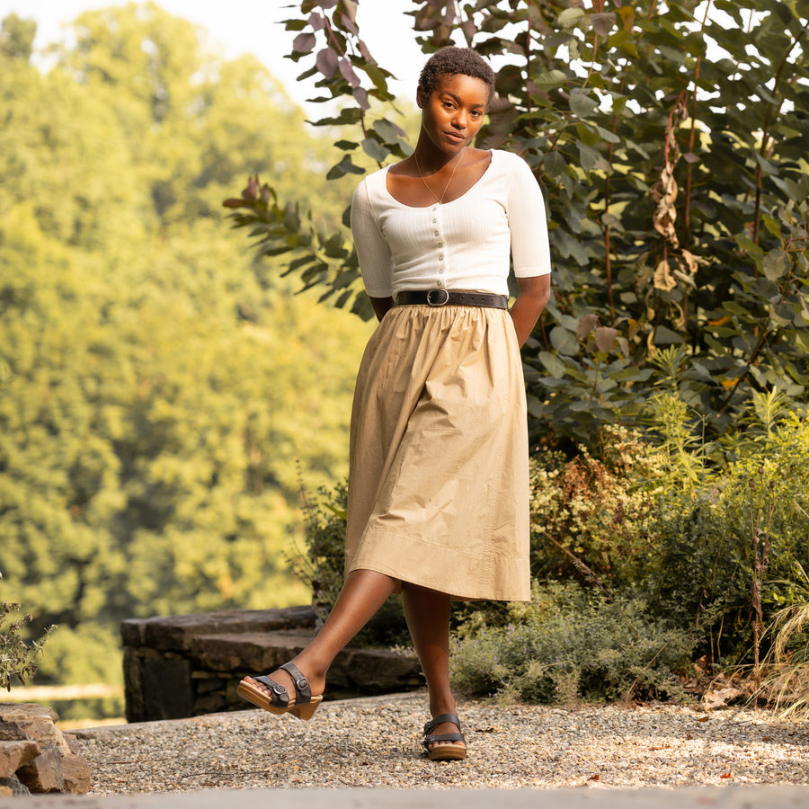 A woman standing in a garden in a white top and tan skirt while wearing black two-strap sandals.