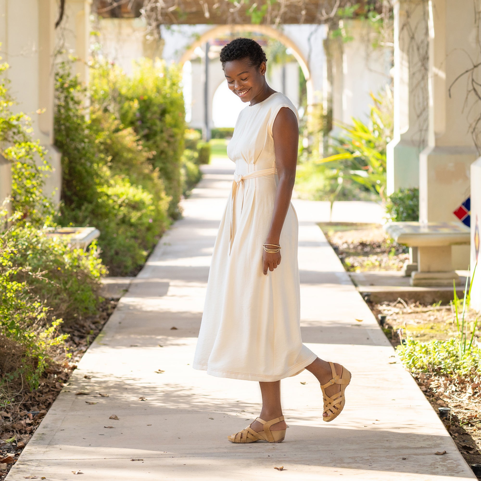 A joyful woman in a flowing white dress standing in a garden and wearing tan fisherman sandals.