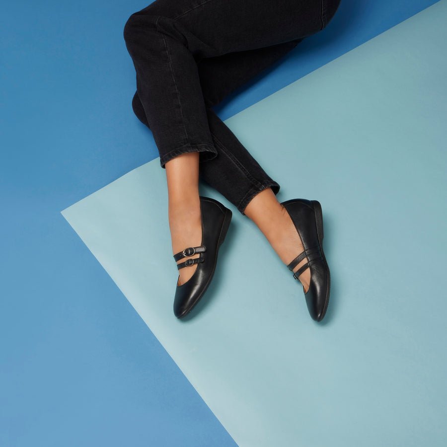A woman laying on a blue background wearing black Mary Janes with a stylish two-strap design.